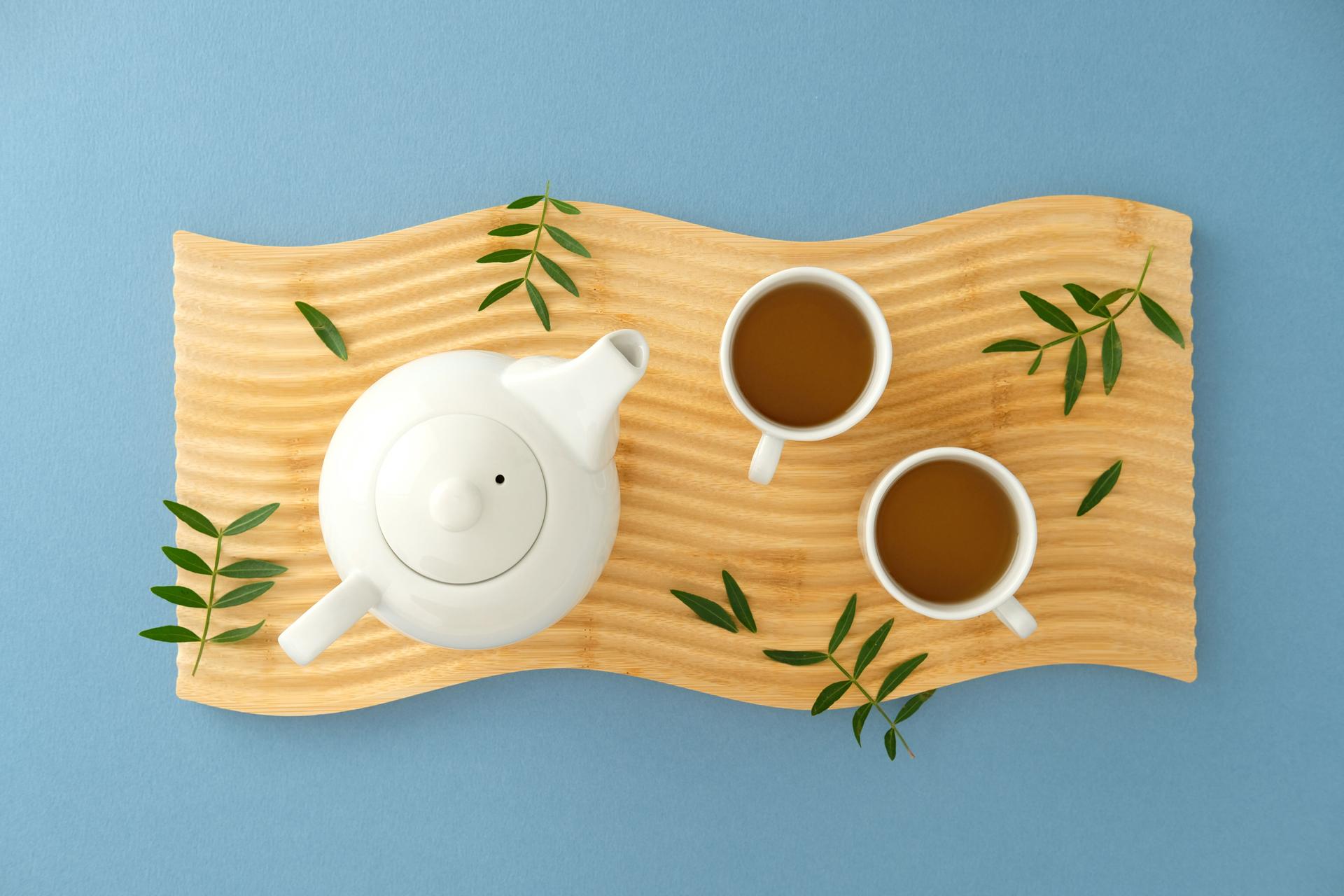 White teapot and cups on wavy wooden tray over blue background, accented with green leaves for a fresh, modern style.
